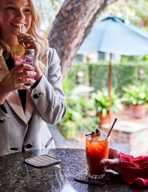 Two people having drinks at a table outside with plants and an umbrella in the background, enjoying a casual conversation.