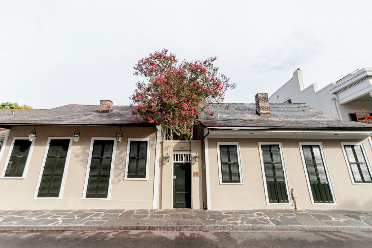 A beige stucco building with green shutters and a gray roof, with a tree in front.