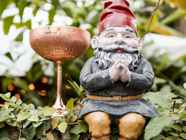 A garden gnome with a red hat is sitting among greenery, next to a bronze birdbath or decorative cup on a stand.