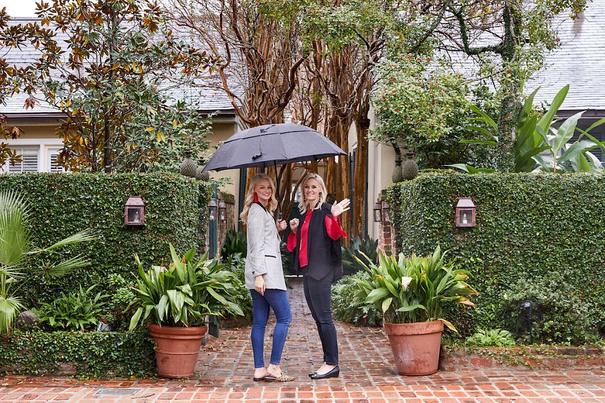 Two people stand on a brick path with an umbrella, surrounded by lush greenery and potted plants, in front of a cozy-looking house.