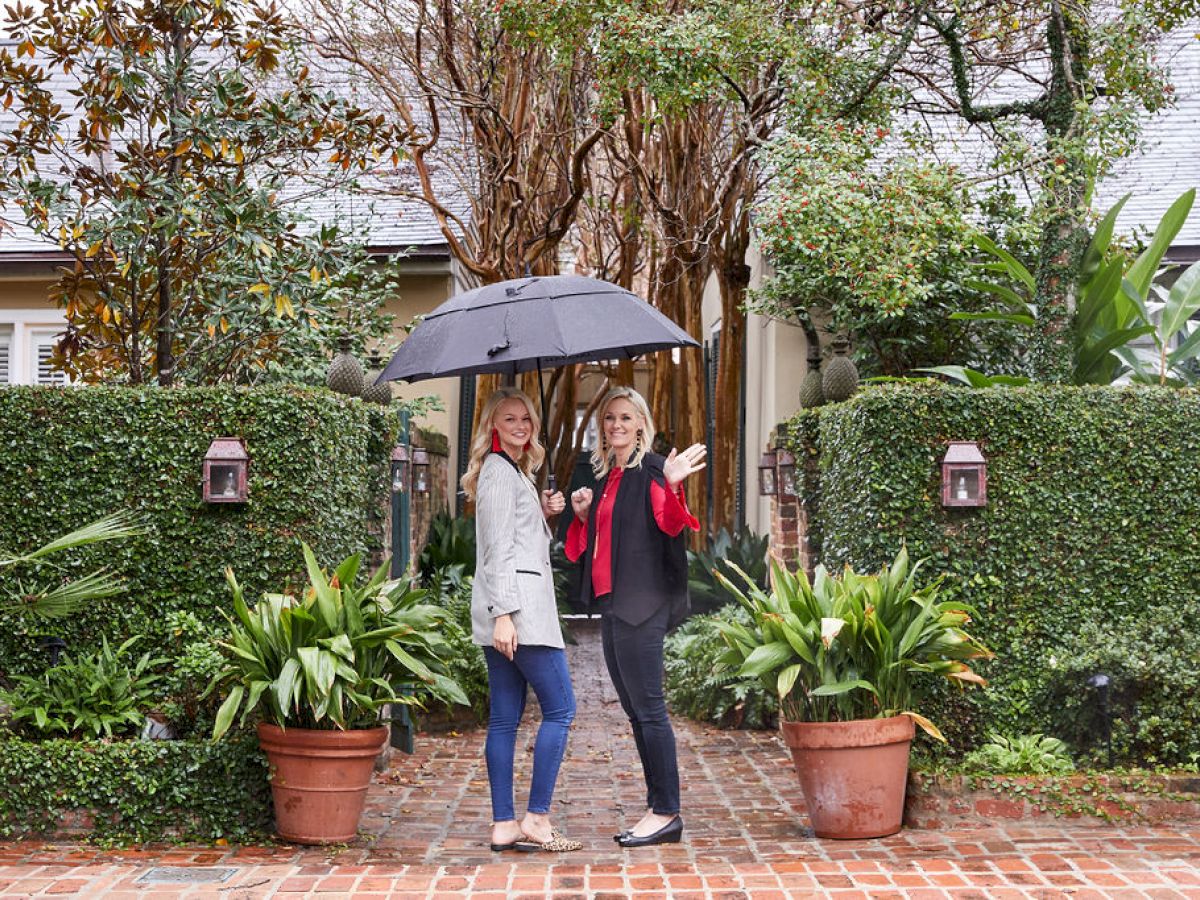 Two people stand on a brick path with an umbrella, surrounded by lush greenery and potted plants, in front of a cozy-looking house.
