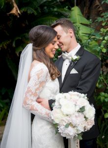 A couple, dressed in wedding attire, embraces lovingly with lush greenery in the background.