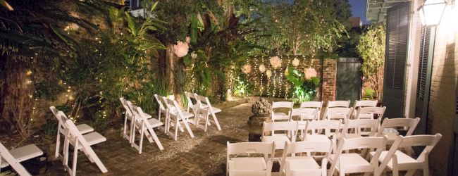 An outdoor setting with white chairs arranged for an event, surrounded by greenery and string lights, under a dimly lit evening sky.
