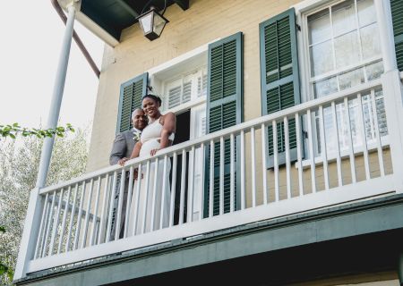 A couple stands on a balcony, smiling, with green shutters and a light-colored building behind them.