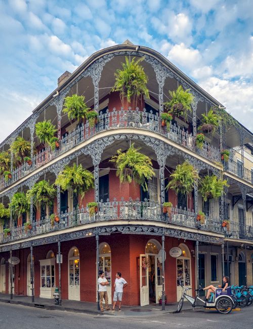 A historic building with ornate balconies covered in plants on a street corner, under a blue sky with clouds.