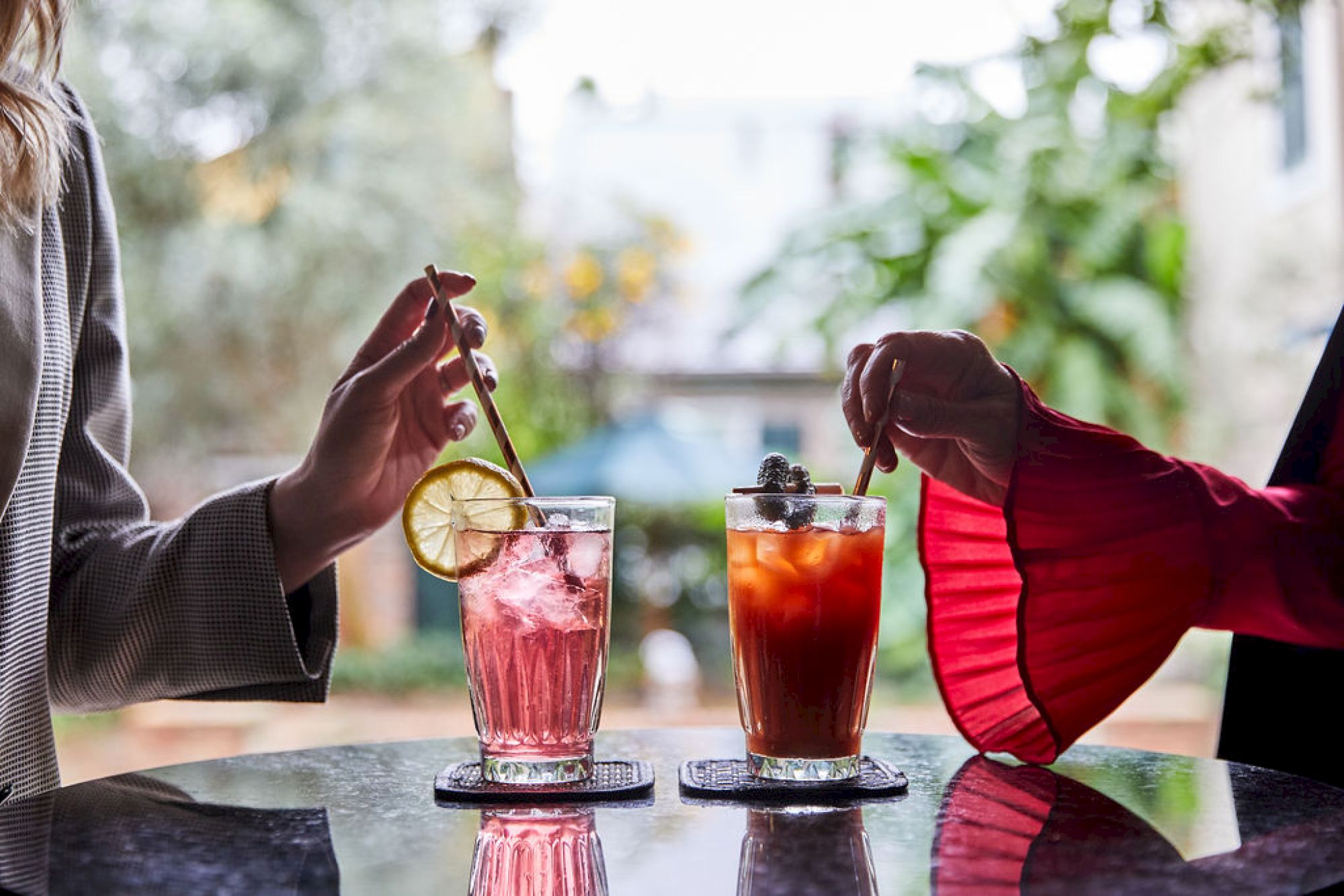 Two people are stirring drinks with garnishes, one with a lemon slice, on a reflective table outdoors.