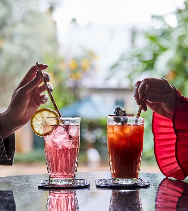 two hands holding straws over glasses of pink and red drinks with ice and garnishes