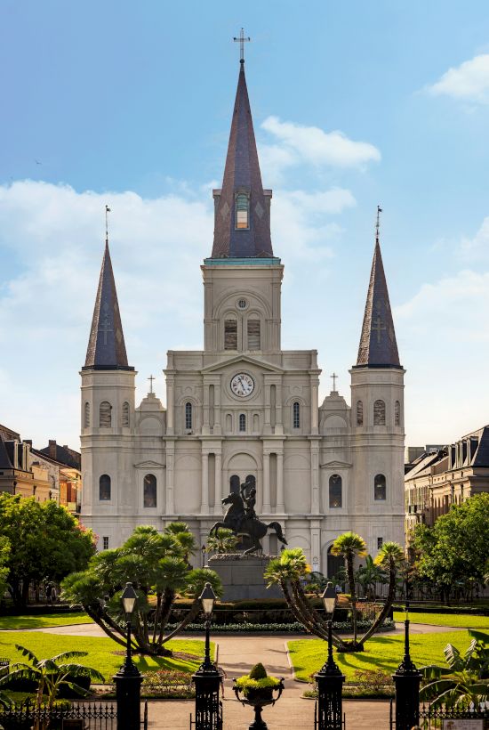 The image shows a grand cathedral with three spires and a statue in front, set in a landscaped park under a clear blue sky.