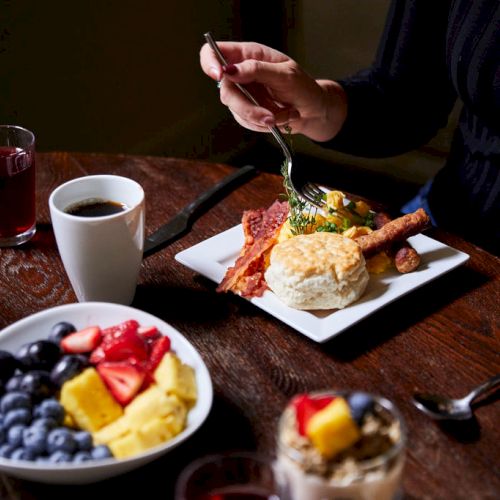 A person is eating breakfast with bacon, eggs, a biscuit, and fruit. Nearby are coffee, juice, and a bowl of assorted berries and yogurt.