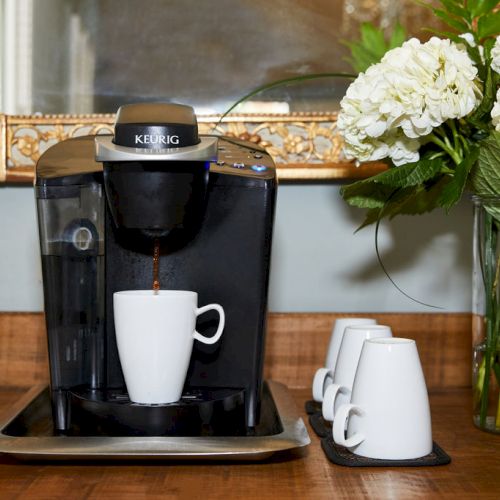 A coffee machine with a mug is brewing coffee. Three additional mugs are nearby, next to a vase with white flowers on a wooden counter.