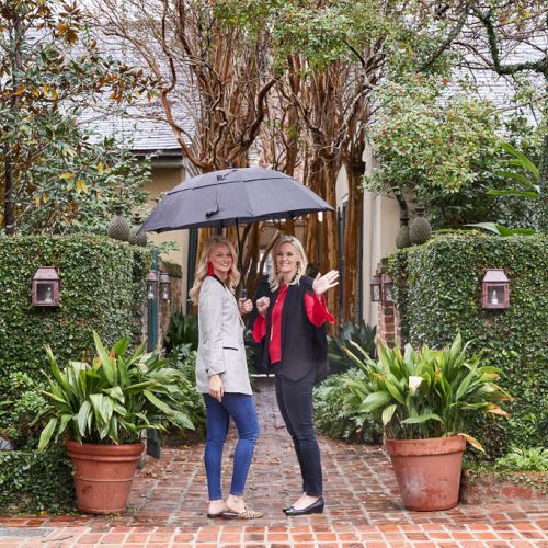 Two people stand on a brick path at a garden entrance, holding an umbrella. Greenery and potted plants surround them on a cloudy day.