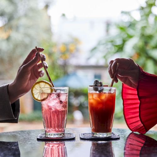 Two people enjoying colorful drinks with garnishes on a table outdoors, blurring into a garden or park background.