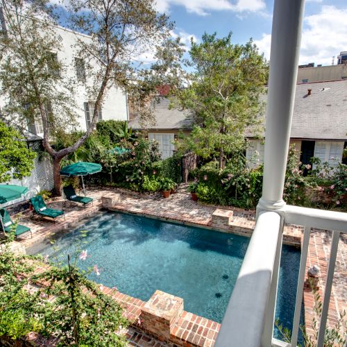 A backyard scene with a rectangular pool, surrounded by greenery, brick patio, and lounge chairs with umbrellas.