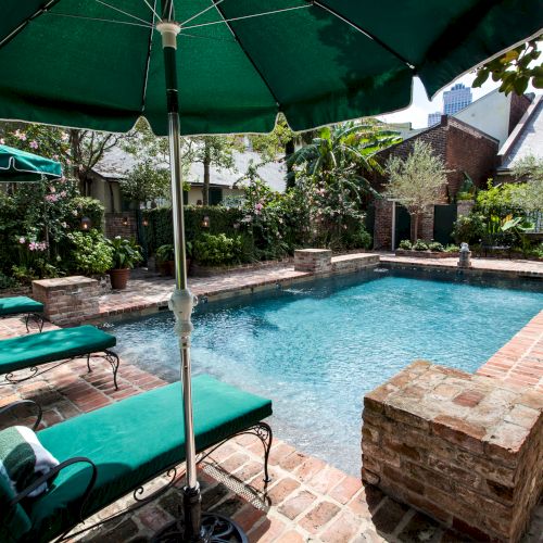 A relaxing pool area with brick surroundings, green loungers, and umbrellas, set amidst lush greenery and nearby buildings.