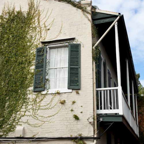 A two-story building with green shutters, a balcony, and ivy growing on white brick walls under a partly cloudy sky.