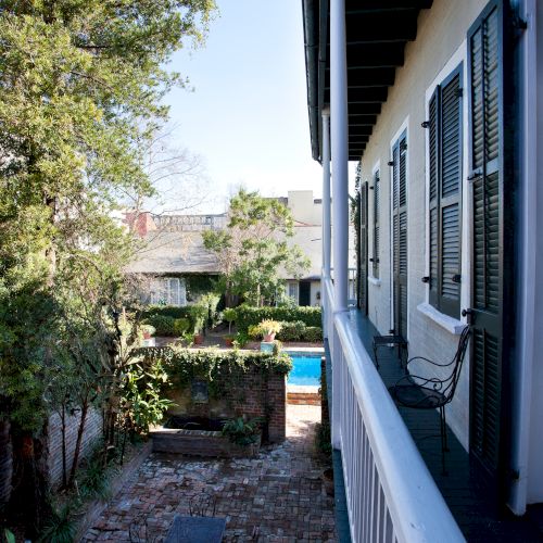 A walkway with brick paving leads past a porch on the right with shutters on windows, alongside trees and a view of a garden and pool.