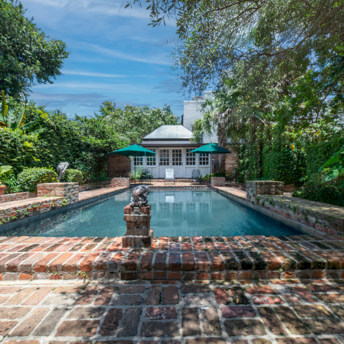 A serene backyard pool surrounded by brickwork and lush greenery, with a small building at one end and clear blue sky above.