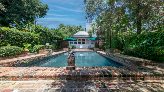 A rectangular pool surrounded by brick patio and lush greenery, with a small building in the background.