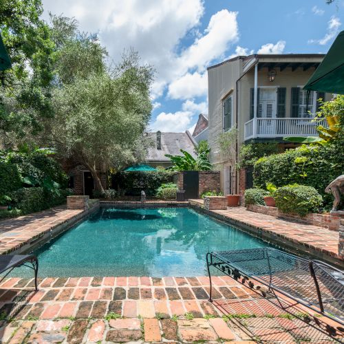 A serene backyard features a rectangular pool, surrounded by lush greenery, brick patio, and lounge chairs under umbrellas.