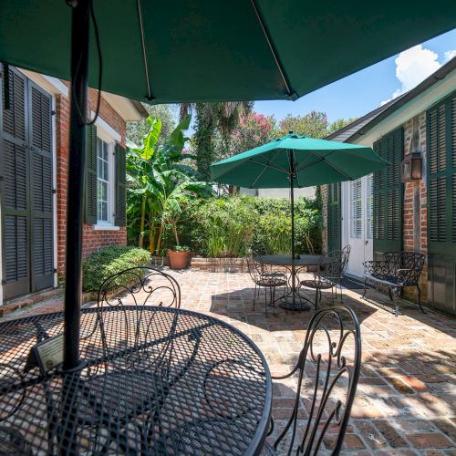 A charming patio features iron tables, chairs, umbrellas, brick flooring, and lush greenery under a clear sky.