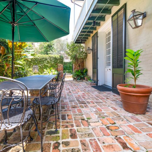 A patio with brick flooring, a metal table and chairs, a potted plant, and a large umbrella is shown beside a building with shuttered windows.