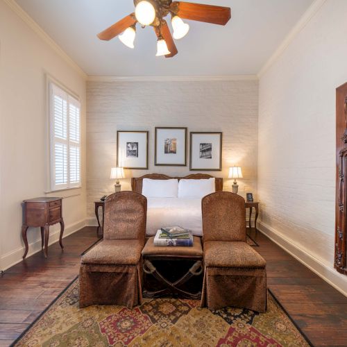 A cozy bedroom with a bed, armchairs, ornate mirror, ceiling fan, rug, and framed pictures on the wall, creating a warm atmosphere.