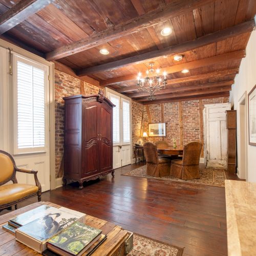 The image shows a rustic room with wooden ceiling beams, a brick wall, vintage furniture, and a chandelier, giving a cozy and classic vibe.