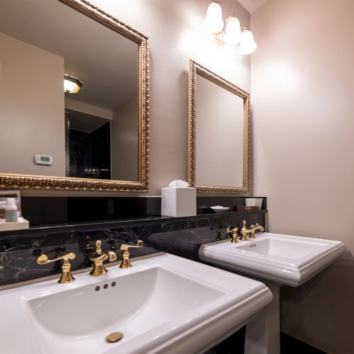 The image shows a bathroom with two sinks, gold fixtures, two mirrors, and overhead lighting against a beige wall.