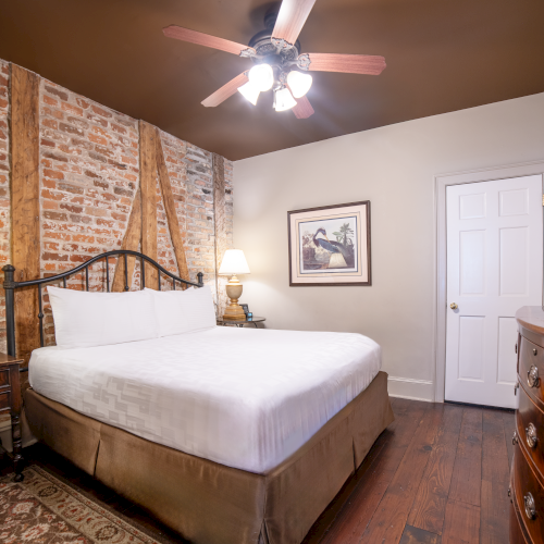 A cozy bedroom with a brick accent wall, wooden beams, a bed, lamps, artwork, dresser, TV, and ceiling fan, creating a warm atmosphere.
