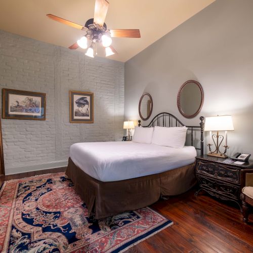 A cozy bedroom with a large bed, two lamps, a ceiling fan, and artwork on the wall; a TV is on the dresser next to a chair.