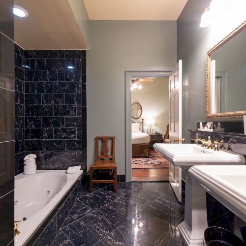 A luxurious bathroom with dark tiles, a bathtub, dual sinks, and a mirror. A wooden chair is near the doorway to the adjoining room.