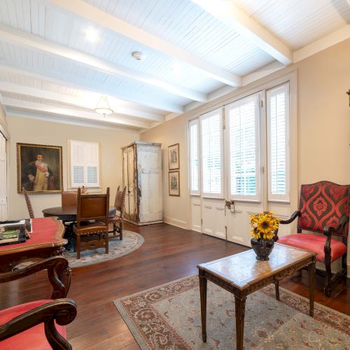 A stylish living room with antique furniture, a grandfather clock, a wall painting, and a television, illuminated by natural light.