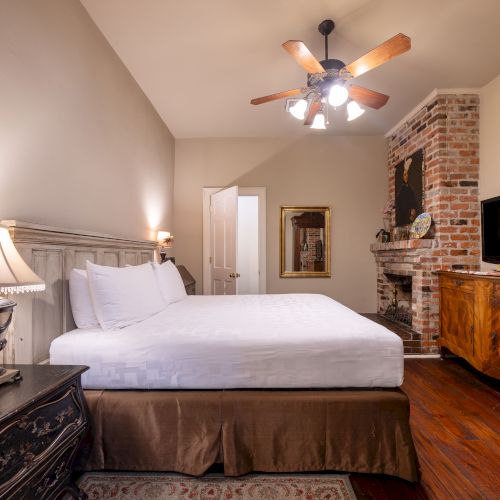 A cozy bedroom with a large bed, side tables with lamps, TV on a dresser, ceiling fan, and a brick fireplace, creating a warm ambiance.