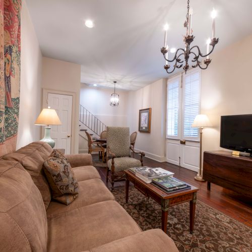 A cozy living room with a brown couch, TV, chandelier, and artwork on the wall, featuring a staircase in the background.