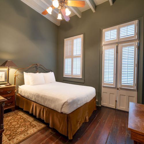 A cozy bedroom with a bed, nightstand, lamp, wooden floor, TV, ceiling fan, and window with shutters.