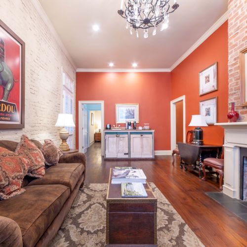 A cozy living room with a brown sofa, colorful art, fireplace, and wooden floors, featuring a chandelier and red accent wall.
