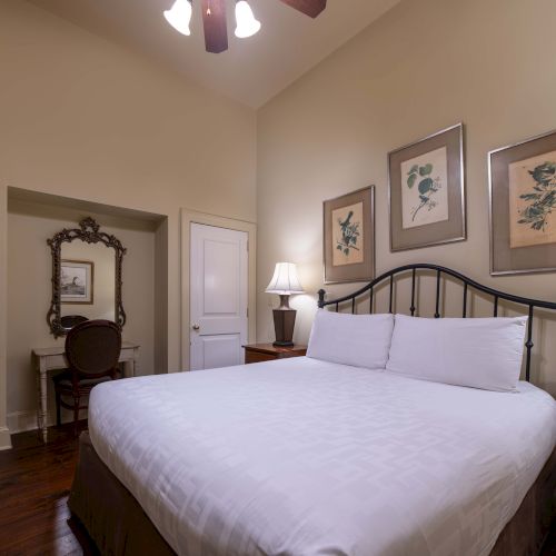 A cozy bedroom features a large bed, elegant lamps, framed botanical prints, a vintage mirror, and a desk with a chair, creating a warm ambiance.