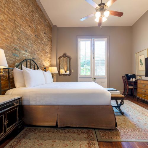 A cozy bedroom with a double bed, brick accent wall, lamps, TV, ceiling fan, dresser, and rug, featuring a serene and inviting atmosphere.