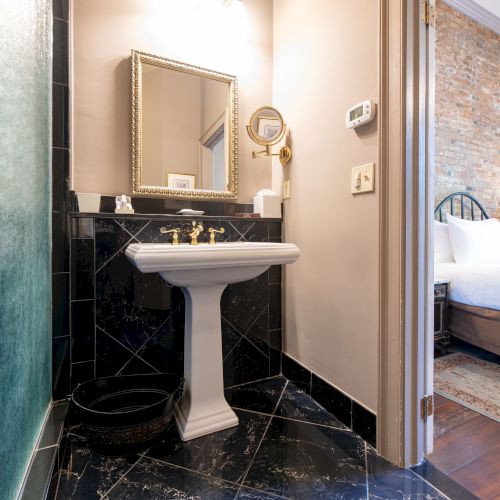 The image shows a bathroom with black tiles, a pedestal sink, and a framed mirror, adjacent to a bedroom with a brick wall and bed.