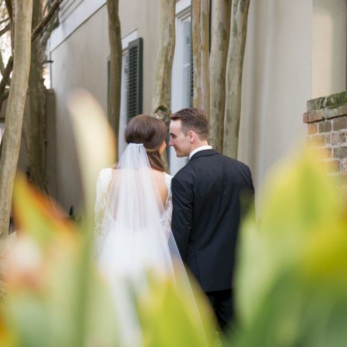 A bride and groom share a moment in a garden setting, surrounded by greenery and a brick wall, with the bride in a veil and gown.