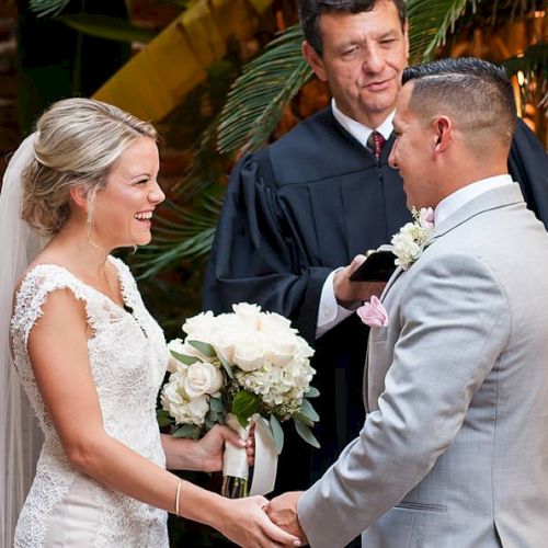 A bride and groom hold hands, smiling at each other during a wedding ceremony, with an officiant standing behind them.