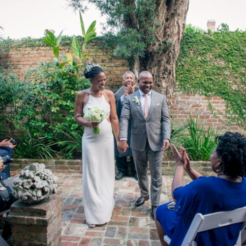 A couple is walking down an aisle at an outdoor wedding ceremony, with guests clapping and smiling.