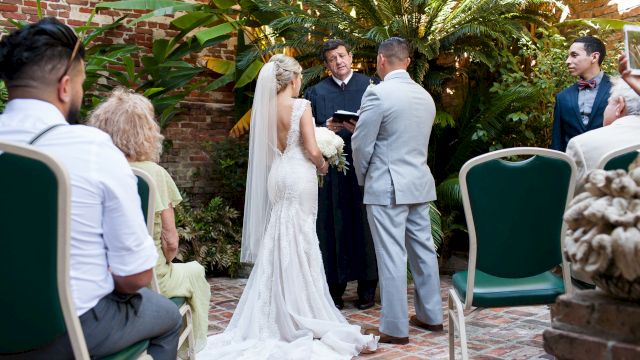 A wedding ceremony taking place in an outdoor setting with a brick patio, green chairs, and plants.
