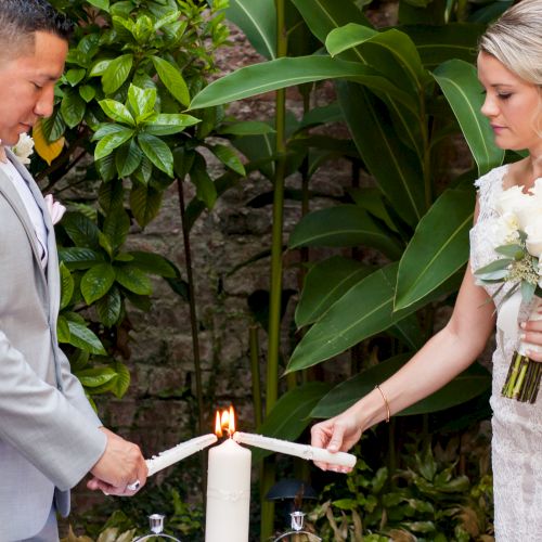 A bride and groom lighting a unity candle together during their wedding ceremony outdoors, surrounded by greenery.