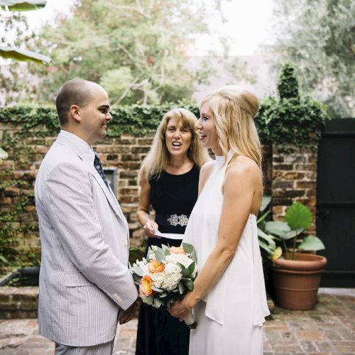 A couple is getting married in a garden setting, with a woman officiating. The bride holds a bouquet of flowers, and they are all smiling.