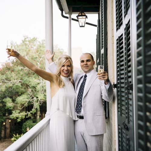 A couple stands on a balcony, celebrating with raised champagne glasses and smiling, surrounded by greenery and a building.