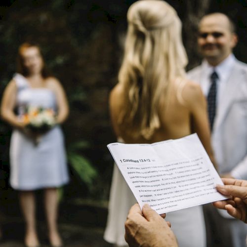 A person holds a paper with text, likely a script, at a wedding ceremony with a couple standing in the background.