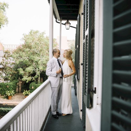 A couple stands on a balcony surrounded by greenery, with the man in a suit and the woman in a white dress, sharing an intimate moment.