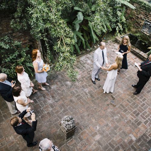 A small outdoor wedding ceremony with a couple getting married, officiant, and guests in a garden with brick pathways and greenery.