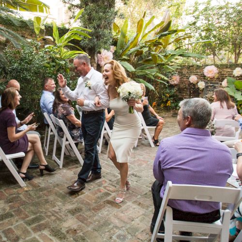 A couple joyfully exits a small outdoor wedding ceremony, surrounded by seated guests on white chairs.